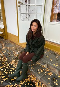 Donna con i capelli lunghi e scuri che indossa un cappotto verde e stivali di gomma abbinati, seduta su gradini di pietra coperti di foglie autunnali.