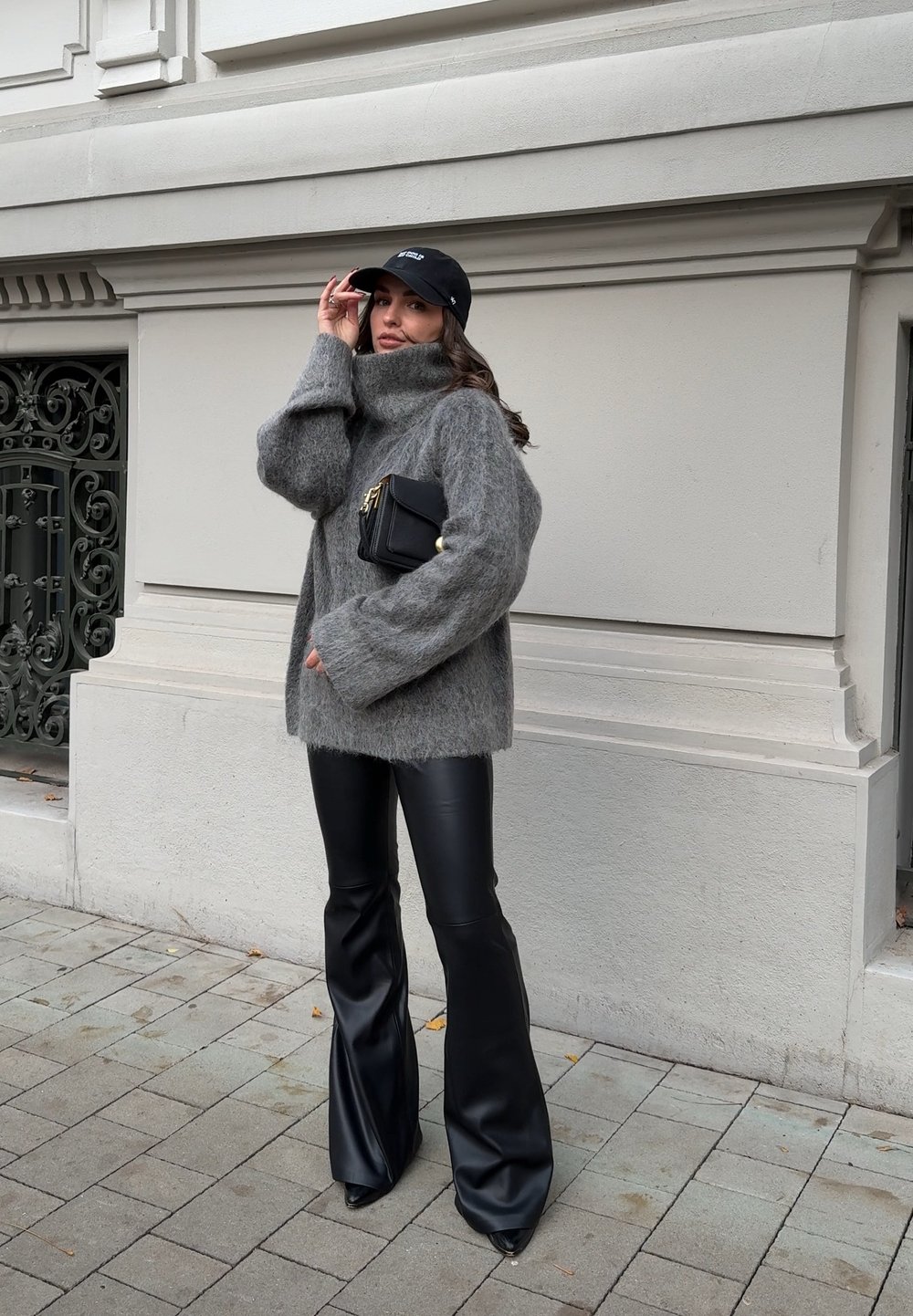 Woman in a grey fuzzy jumper and black flared leather trousers adjusting her black cap while holding a black handbag, standing on a city pavement.