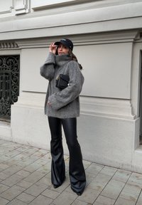 Woman in a grey fuzzy jumper and black flared leather trousers adjusting her black cap while holding a black handbag, standing on a city pavement.