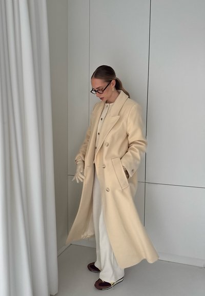 Abrigo de lana crema de doble botonadura con solapa, usado sobre una blusa clara, combinado con pantalones crema y zapatos oscuros. Textura suave, ajuste entallado.