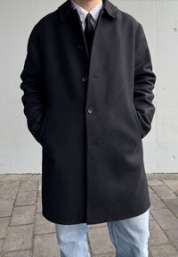 Manteau en laine noire mélangée avec un col, fermeture à boutons et poches latérales. Assorti à un jean bleu clair et une chemise rayée en dessous.