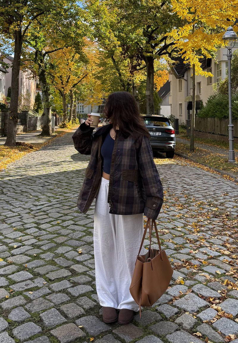 Brown checked jacket, dark top, white wide-leg trousers, brown bag, holding a coffee cup. Stone pavement and trees with autumn foliage in the background.
