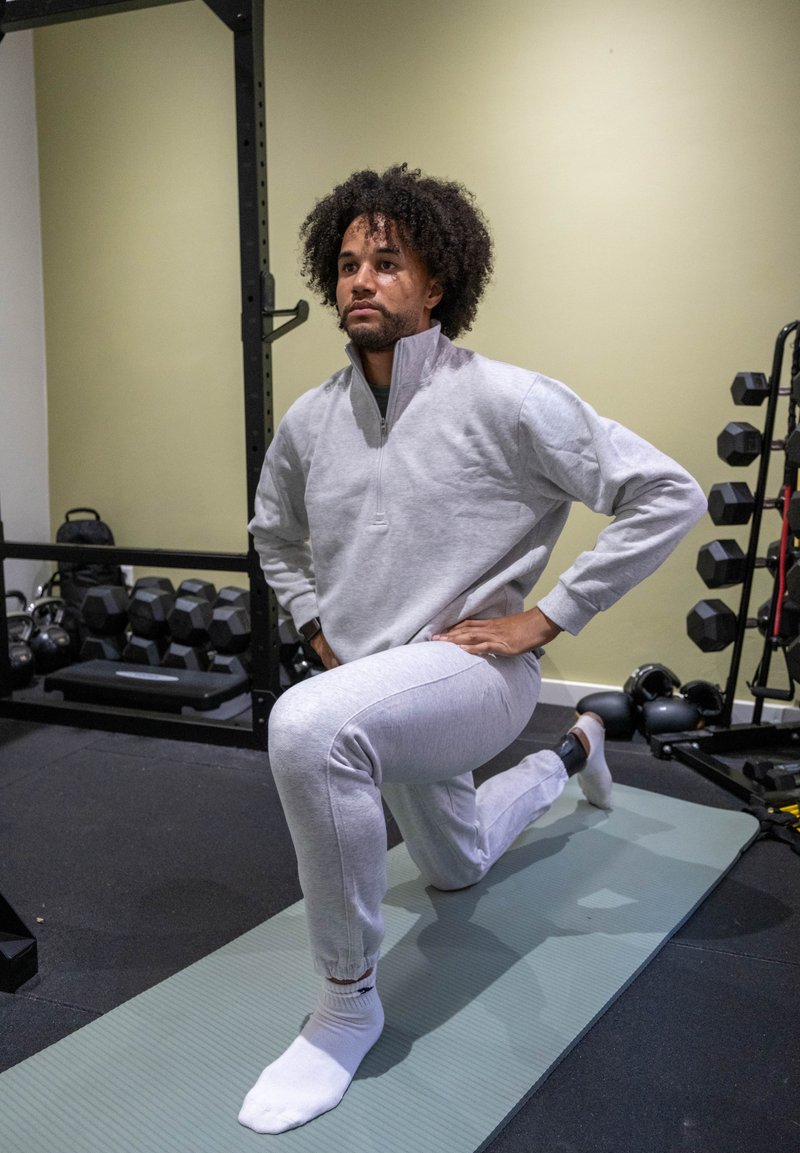 Conjunto desportivo cinza claro com uma camisola de fecho de correr até ao peito e calças afuniladas combinando. Indivíduo realiza um avanço numa esteira de exercício verde num ginásio.