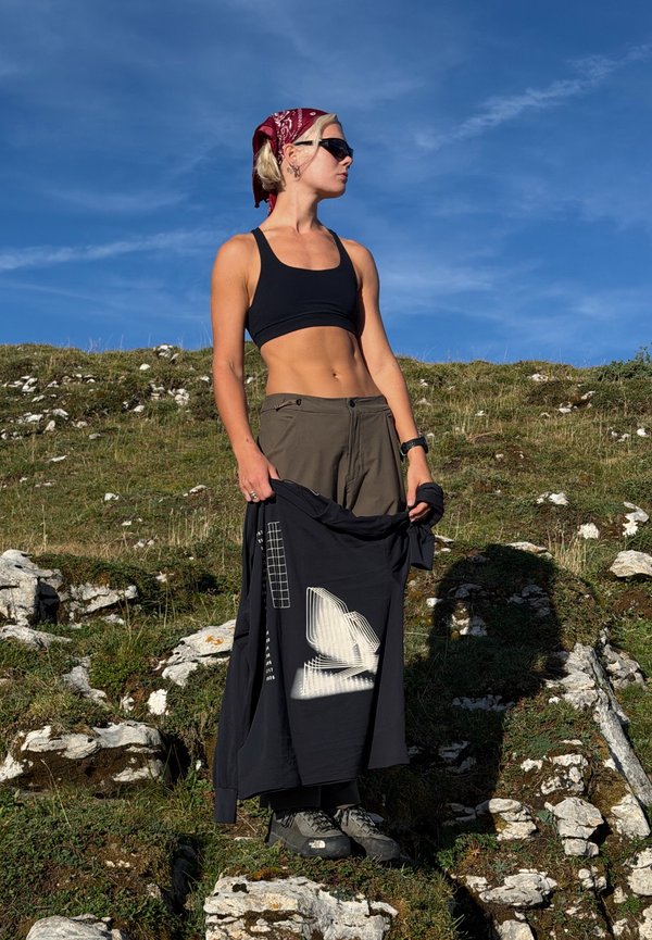 Donna con occhiali da sole, reggiseno sportivo, bandana e scarpe da trekking in piedi su una collina rocciosa che tiene una giacca nera sotto un cielo azzurro limpido.