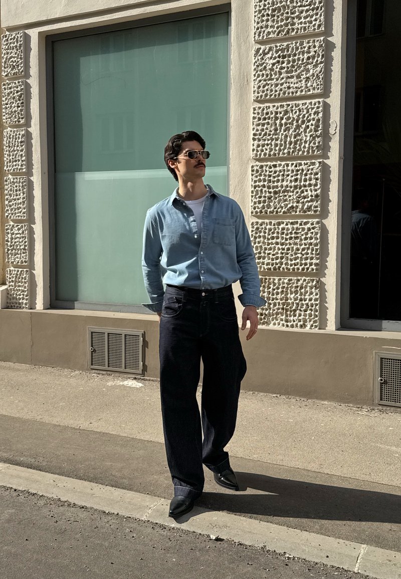 Homme portant des lunettes de soleil, une chemise bleu clair, un pantalon large sombre et des chaussures noires marchant sur un trottoir ensoleillé près d'un mur de bâtiment texturé.