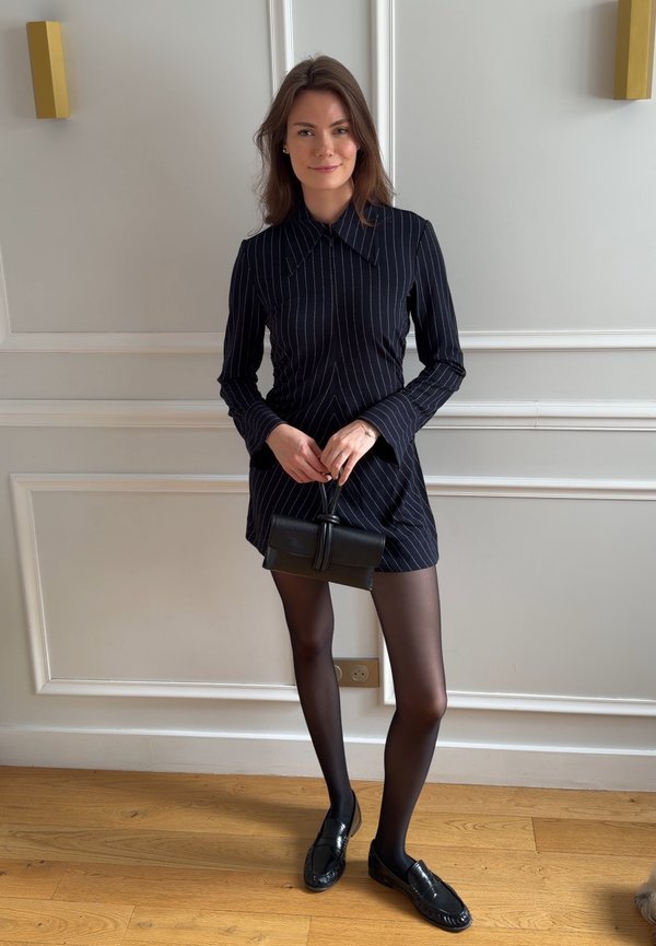 Frau trägt ein schwarzes Kleid mit Nadelstreifen, transparente schwarze Strumpfhosen und schwarze Loafers, hält eine kleine schwarze Handtasche, steht drinnen auf Holzboden.