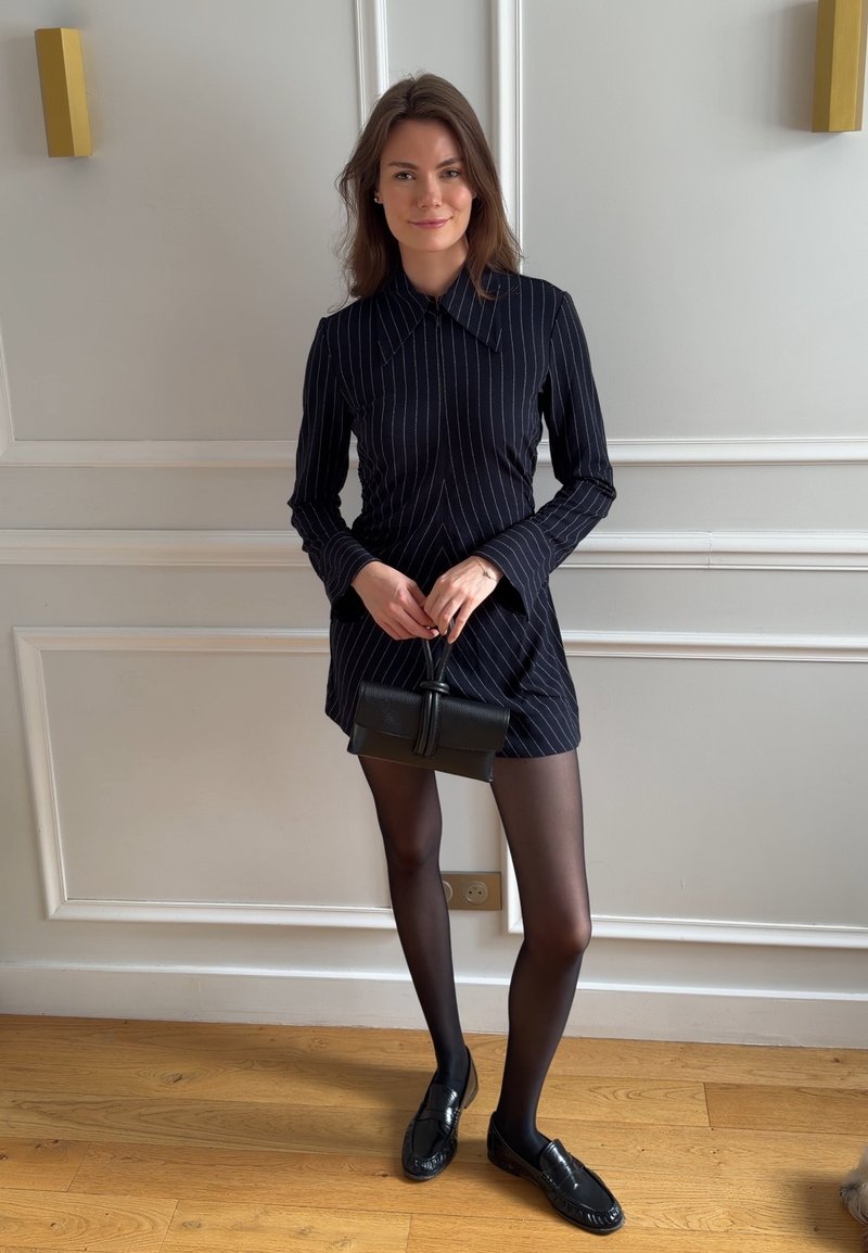 Femme portant une robe noire à fines rayures, des collants noirs transparents et des mocassins noirs, tenant un petit sac à main noir, debout à l'intérieur sur un sol en bois.