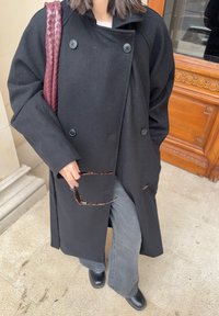 Dlhý čierny vlnený kabát s dvojradovými gombíkmi, nosený cez sivé nohavice s širokými nohavicami, drží hnedé slnečné okuliare s korytnačím vzorom a prepletenú bordovú tašku.