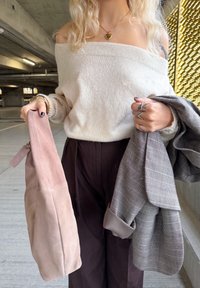Roomwitte off-shoulder trui, donkere wijde broek, houdt een blushroze suède tas en een grijze geruite jas vast. Textuur van stoffen zichtbaar.