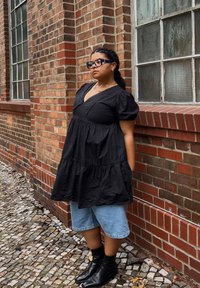 Vestido preto de mangas bufantes com decote em V e design de camadas de babados, combinado com calções de ganga azul-claro e botas pretas de tornozelo.