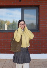Jonge vrouw met bril, gekleed in een gele jas en een geruite rok, die een grote bruine tas draagt, staande voor een bakstenen muur met ramen.