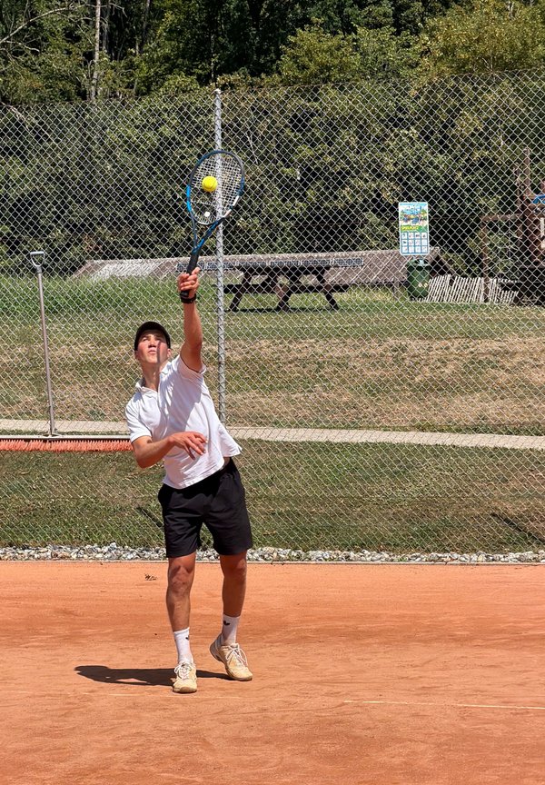 Jeune homme en chemise blanche et short noir servant une balle de tennis sur un court en terre battue extérieure avec une clôture grillagée et des arbres derrière.