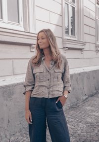 Lniana koszula zapinana na guziki w jasnobrązowym kolorze, z dwoma kieszeniami z przodu, zestawiona z szerokimi ciemnymi spodniami, na tle teksturowanej ściany.