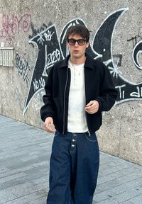 Sort lynlåsjakke, hvid tekstureret henley, oversize blå denimbukser med knapdetalje. Person bærer sorte briller og ringe. Urban baggrund.