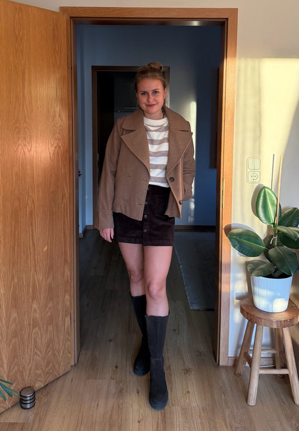Une femme se tient dans l’encadrement d’une porte, vêtue d’une veste brune, d’un pull rayé, d’une jupe noire et de bottes montant jusqu’aux genoux. Une plante en pot se trouve à côté d’elle.