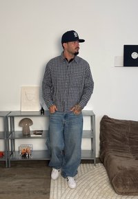 Chemise à carreaux boutonnée en gris et marine, portée avec un jean bleu ample. Des baskets blanches complètent la tenue, posée dans un cadre intérieur simple.