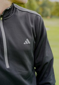 Sivi pulover z zadrgo do polovice iz teksturirane tkanine s kontrastno svetlo sivo zadrgo, z logotipom Adidas na levi strani prsnega koša.
