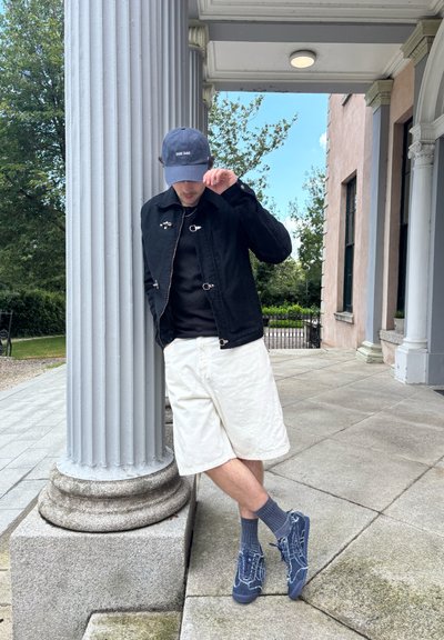 Chaqueta negra de botones, camiseta negra, pantalones cortos blancos, gorra azul marino con "PAPÁ PERRO", calcetas grises y zapatos azul marino con detalles texturizados. De pie junto a una columna.