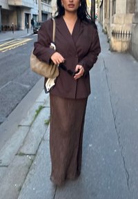 Smeđa oversized sako nosi se preko smeđe teksturirane maksi suknje, u kombinaciji s svijetlosmeđom torbom i držeći sunčane naočale. Urbani ambijent.