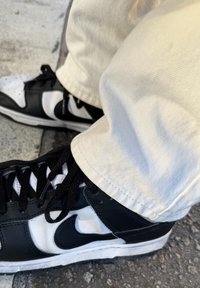 Black and white Nike trainers feature a textured leather upper, a prominent logo, and contrasting laces. Light beige trousers with a slight cuff.