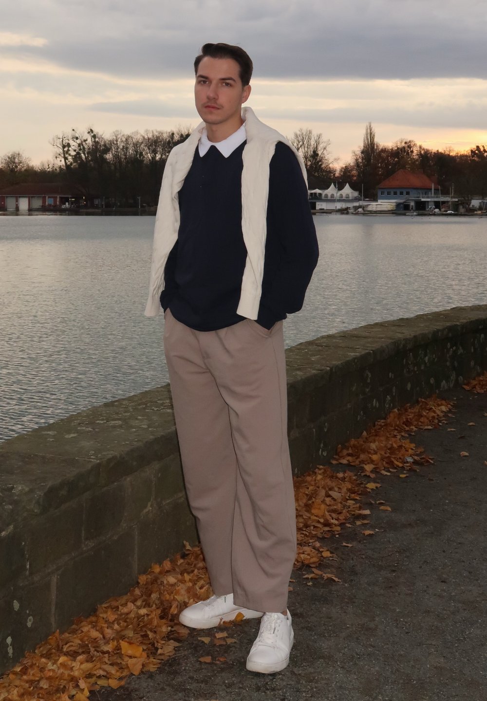 Junger Mann, der einen marineblauen Pullover, eine cremefarbene Weste, beige Hosen und weiße Turnschuhe trägt, steht an einer Steinmauer neben Wasser mit herbstlichen Blättern.