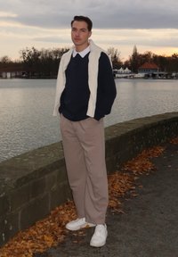 Junger Mann, der einen marineblauen Pullover, eine cremefarbene Weste, beige Hosen und weiße Turnschuhe trägt, steht an einer Steinmauer neben Wasser mit herbstlichen Blättern.