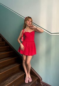 Czerwona sukienka bez rękawów z guzikami i rozkloszowanym dołem. Połączona z różowymi sandałami ozdobionymi różami oraz dopasowaną małą torebką. Tło stanowi drewniane schody.