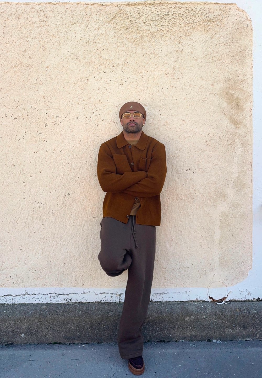 Brown wool cardigan over a beige shirt, teamed with dark brown relaxed trousers and brown shoes. Worn with a matching beanie and glasses.