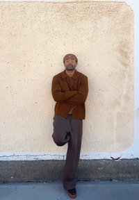 Brown wool cardigan over a beige shirt, teamed with dark brown relaxed trousers and brown shoes. Worn with a matching beanie and glasses.