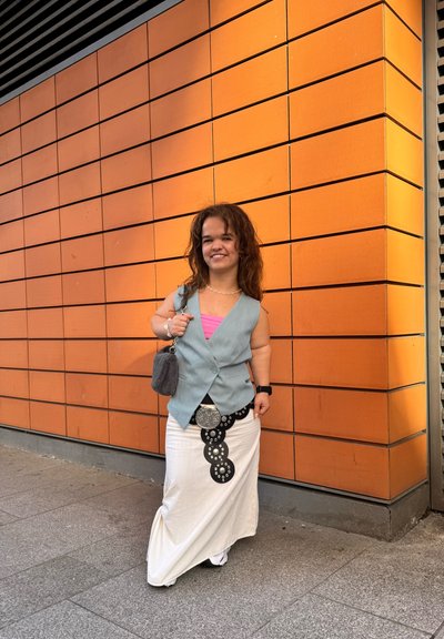 Chaleco sin mangas de color gris claro sobre una blusa rosa, falda blanca drapeada con acentos circulares negros, de pie frente a una pared de azulejos naranjas.