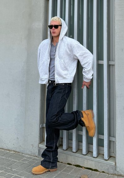 Sudadera gris con cremallera sobre una camiseta gris, jeans de mezclilla oscura y botas marrones. La persona está apoyada contra una pared con listones metálicos verticales.