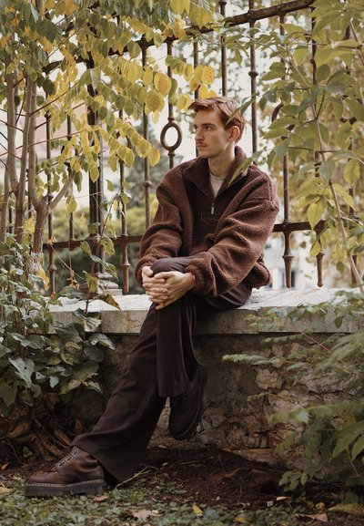 Chaqueta de forro marrón con cremallera, pantalones oscuros y zapatos marrones gruesos. Sentado contra una pared de piedra, rodeado de vegetación y una barandilla de hierro.