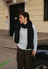 Pull bleu clair tricoté avec un motif texturé, associé à une écharpe noire. Pantalon marron ample, trottoir gris et un véhicule noir.