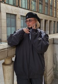 Μαύρο αθλητικό μπουφάν με λευκές ρίγες, χαλαρή εφαρμογή, ελαστικές μανσέτες και μπροστινό φερμουάρ. Φορεμένο με μαύρη μπερέ και υπερμεγέθη γυαλιά ηλίου.