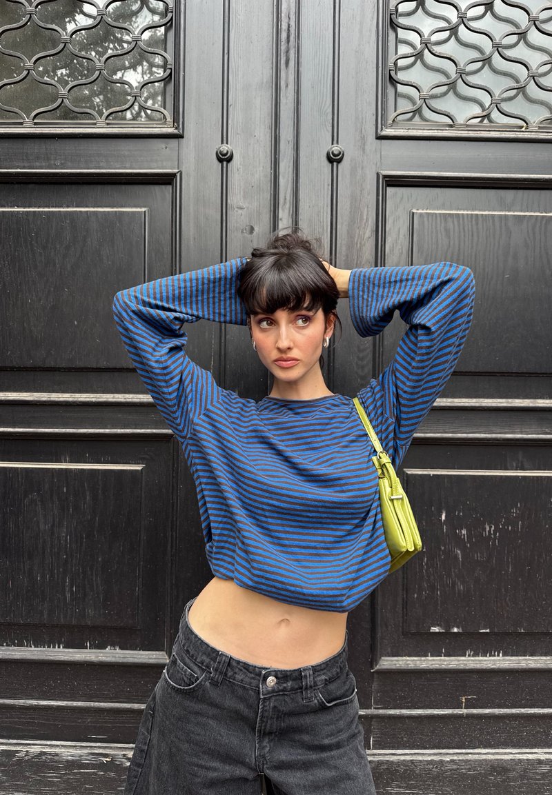 Jeune femme aux cheveux foncés en chignon portant un haut rayé bleu et noir à manches longues et un jean noir, debout les mains derrière la tête devant une porte noire.