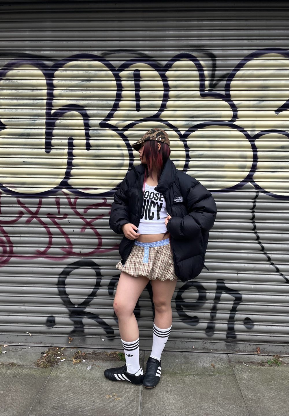 Casaco puffer preto, top cropped branco com texto, saia mini xadrez, meias altas com riscas, sapatos pretos com três riscas, de pé contra um graffiti.