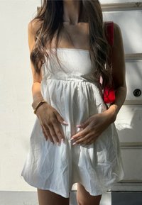 Robe mini blanche avec un design sans bretelles, taille froncée et texture douce. Un sac rouge est porté sur une épaule. Fond clair.