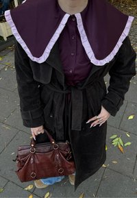 Fekete szarvasbőr trench coat nagy lila gallérral és rózsaszín fodrokkal, lila ing fölött viselve. Barna bőr kézitáskát tart.