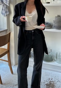 Veste noire portée sur un caraco blanc orné de dentelle, associée à un jean noir taille haute. Tient une tasse en céramique et une petite assiette blanche.