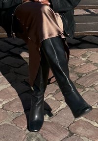 Osoba siedząca na ławce w błyszczącej brązowej spódnicy i czarnych skórzanych butach za kolano, z dłońmi opartymi na kolanach na kostkowej nawierzchni.