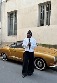 Auto d'epoca dorata con tetto nero, fari tondi e pneumatici con fianco bianco. Persona con camicia bianca, pantaloni neri e berretto che sta accanto ad essa.