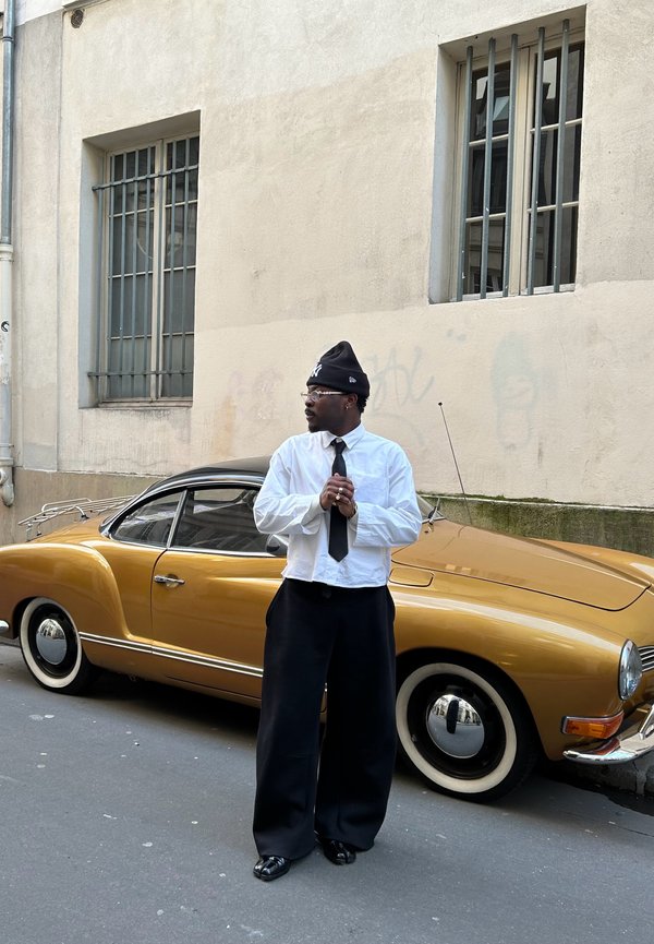 Auto d'epoca dorata con tetto nero, fari tondi e pneumatici con fianco bianco. Persona con camicia bianca, pantaloni neri e berretto che sta accanto ad essa.