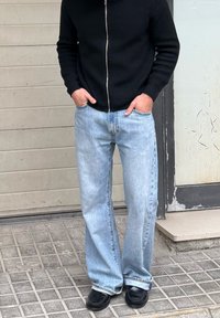 Άτομο που φοράει ανοιχτόχρωμα τζιν, μαύρο φούτερ με φερμουάρ και μαύρα παπούτσια, στέκεται με τα χέρια στις τσέπες πάνω σε πλακόστρωτη επιφάνεια.