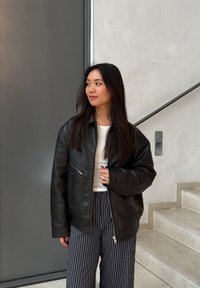 Svart läderbomberjacka med dragkedja fram, sidofickor och avslappnad passform, parat med randiga vida byxor.