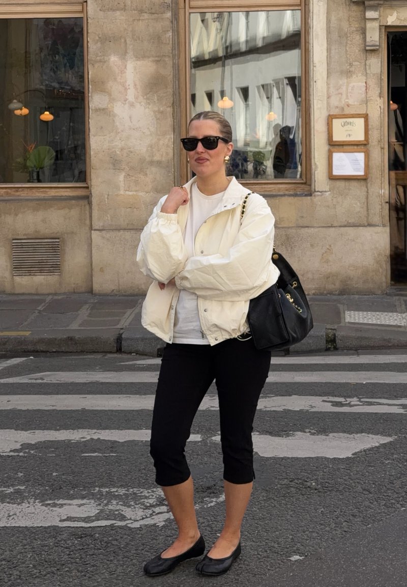 Femme portant des lunettes de soleil, une veste blanche et un pantalon noir, debout sur un passage piéton en ville, tenant un sac à main noir sur son épaule.