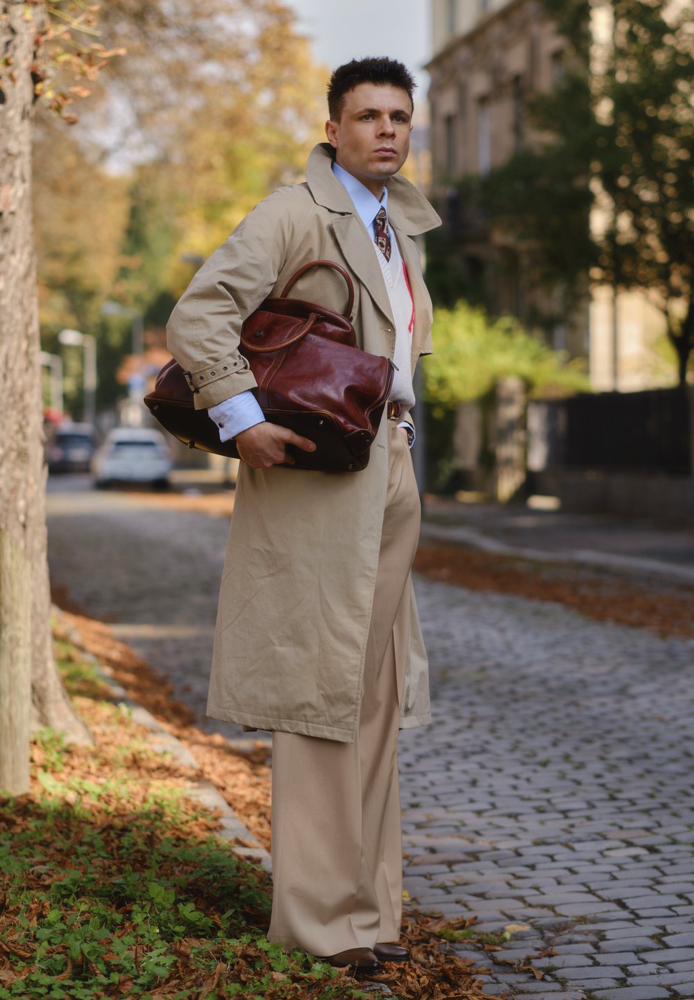Abrigo Trench beige, pantalones ajustados beige, camisa azul, corbata de estampado, bolso de cuero marrón. Entorno natural con hojas de otoño sobre adoquines.