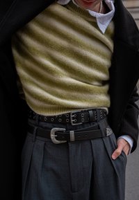 Pull en maille rayé vert et crème superposé sous un manteau noir. Ceinture en cuir noir avec boucle argentée et détails à œillets, portée avec un pantalon gris.
