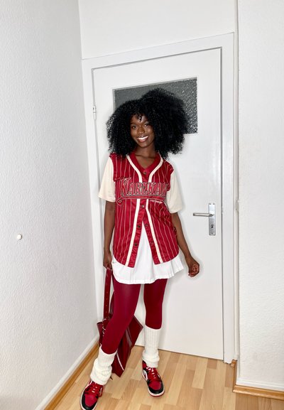 Jersey a rayas rojas y blancas con texto bordado, sobre un vestido blanco, combinado con leggings rojos y calentadores de piernas blancos. Bolso rojo y zapatos.