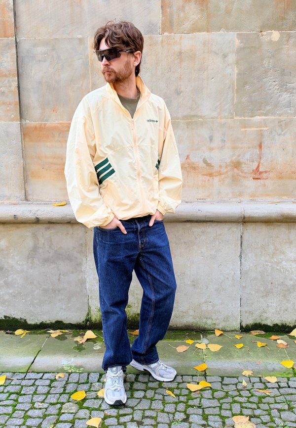 Un homme portant des lunettes de soleil, une veste Adidas couleur crème, un jean et des baskets se tient près d’un mur en pierre, dans une rue pavée jonchée de feuilles.