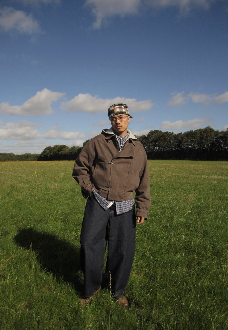 Un hombre con una gorra a cuadros, chaqueta marrón y pantalones oscuros y sueltos está de pie en un campo cubierto de hierba bajo un cielo parcialmente nublado.